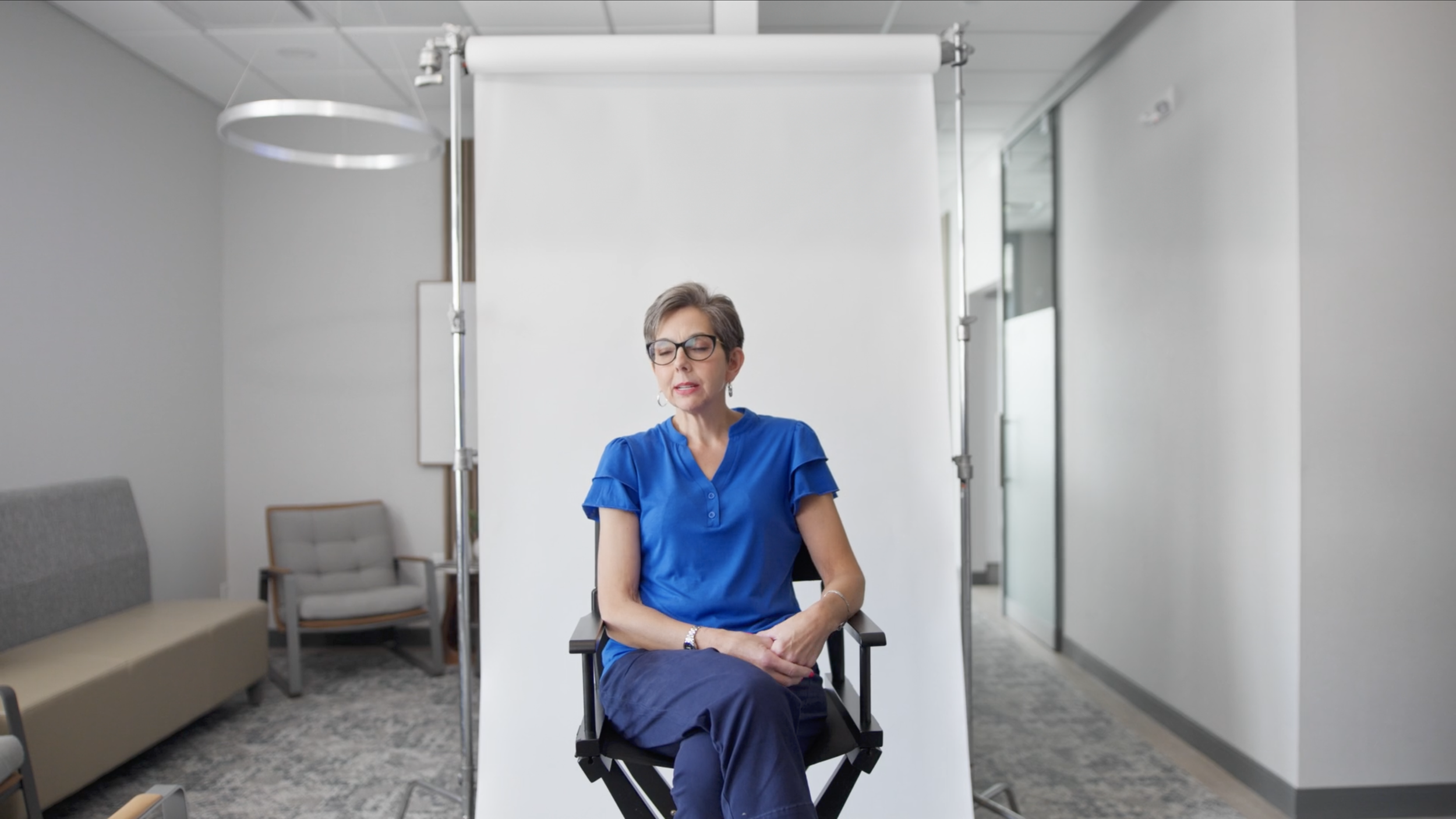 A woman with short gray hair, glasses, and a blue shirt sitting in a director's chair with her eyes closed in a bright, modern office setting, with a white backdrop behind her.