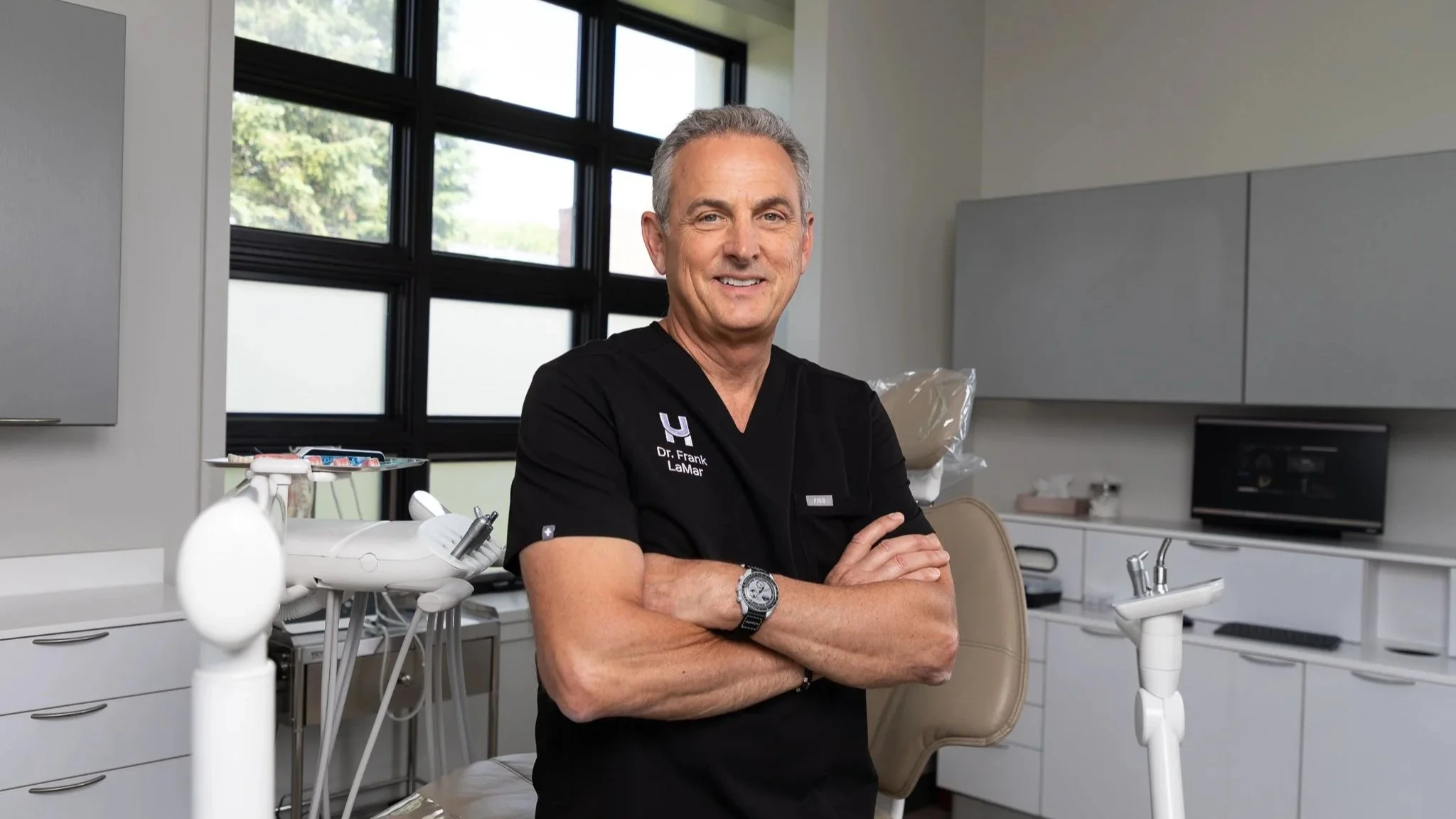 A smiling middle-aged man with gray hair, wearing a black medical uniform with a name tag, standing with arms crossed in a modern dental clinic. The clinic has dental instruments, a dental chair, and cabinets, with large windows in the background.