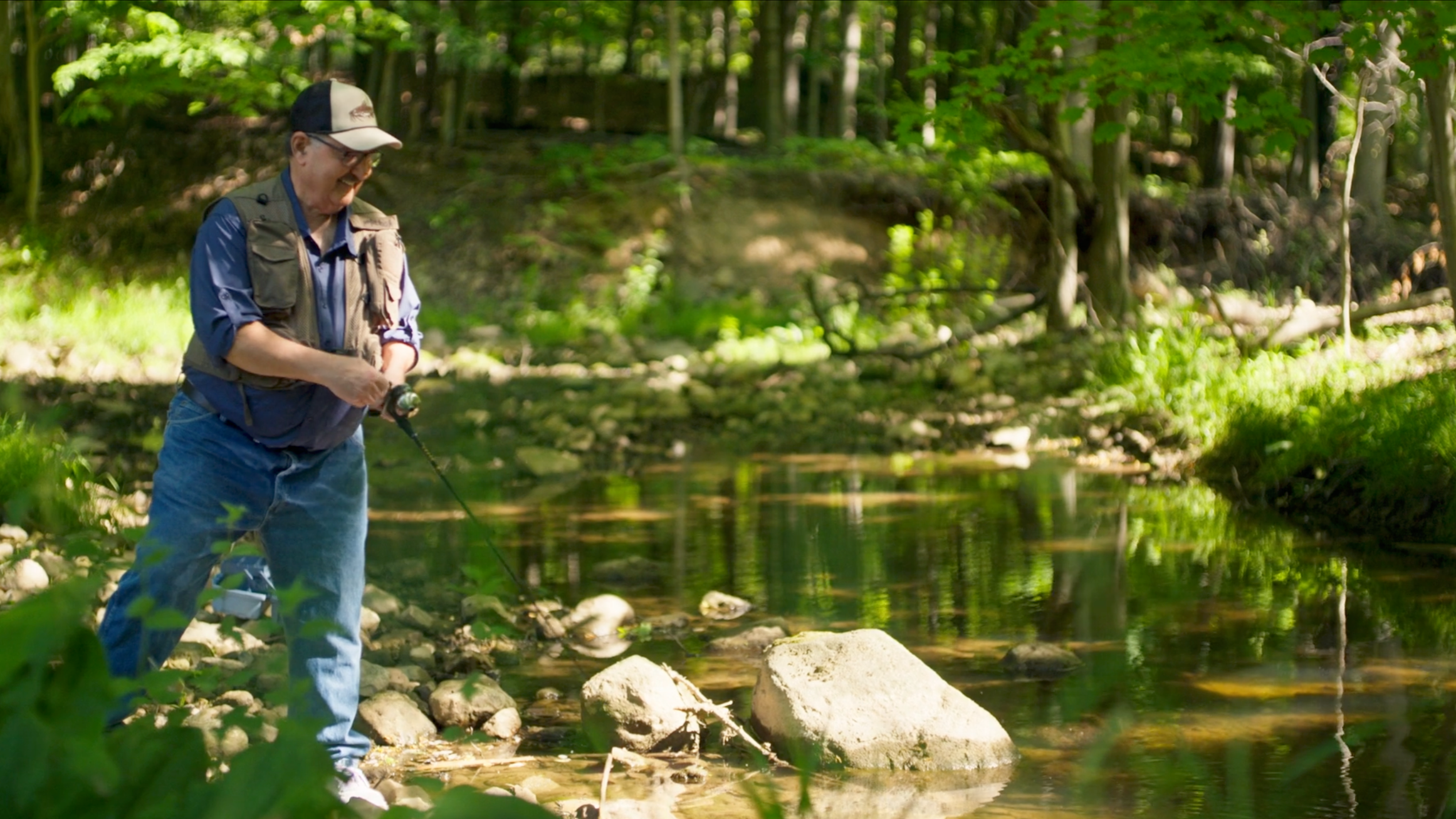 An elderly man wearing a cap, glasses, a beige vest, and blue jeans is fishing in a creek in a wooded area on a sunny day.
