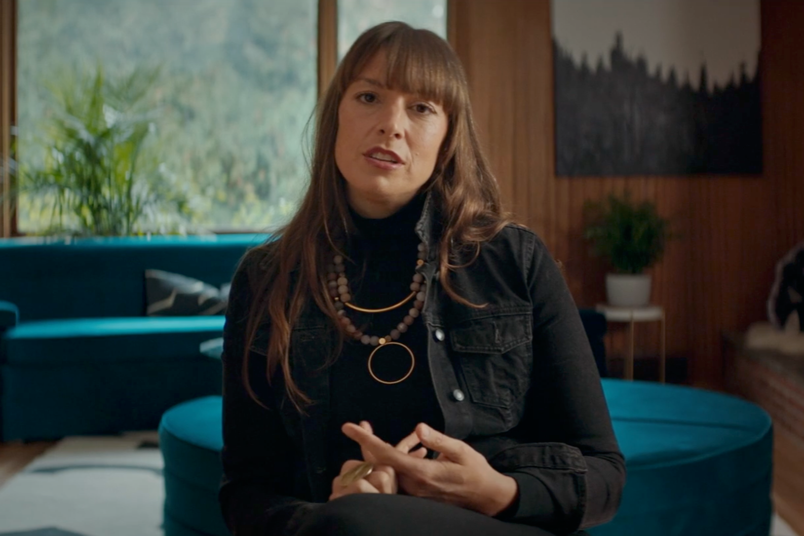 A woman with long brown hair and bangs wearing black clothing and layered necklaces sitting in a living room with large windows, a blue couch, and a wooden wall with potted plants and artwork.