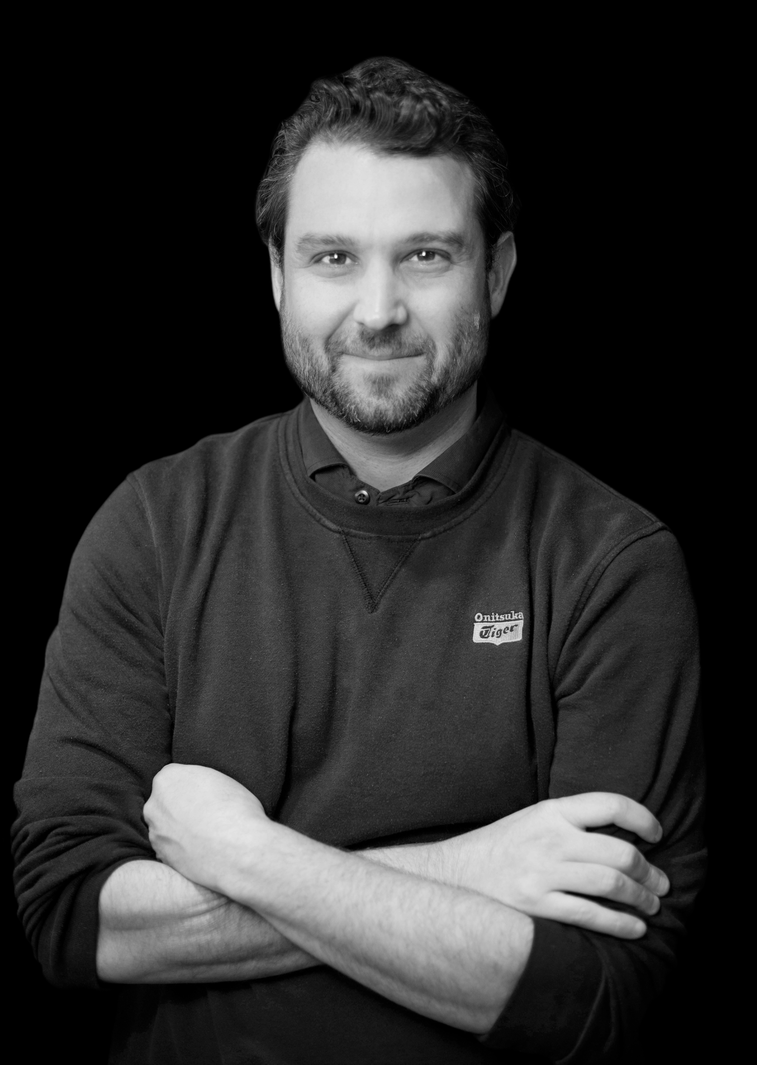 Black and white portrait of a man with short dark hair and a beard, smiling slightly, wearing a dark crew neck sweatshirt with a small logo on the chest, crossing his arms, against a black background.