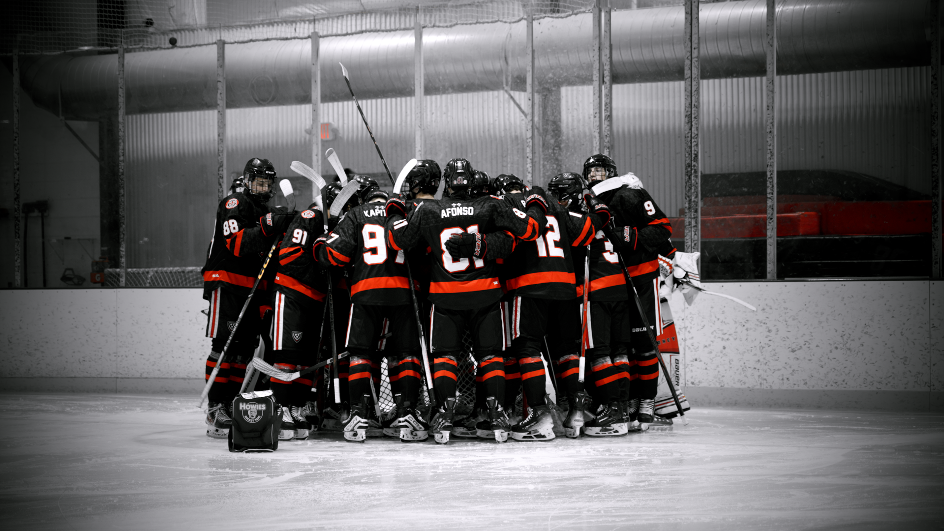 Hockey team in black and red uniforms huddled together on ice rink with ice skate bags nearby, in a mostly black and white photo with some color accents.