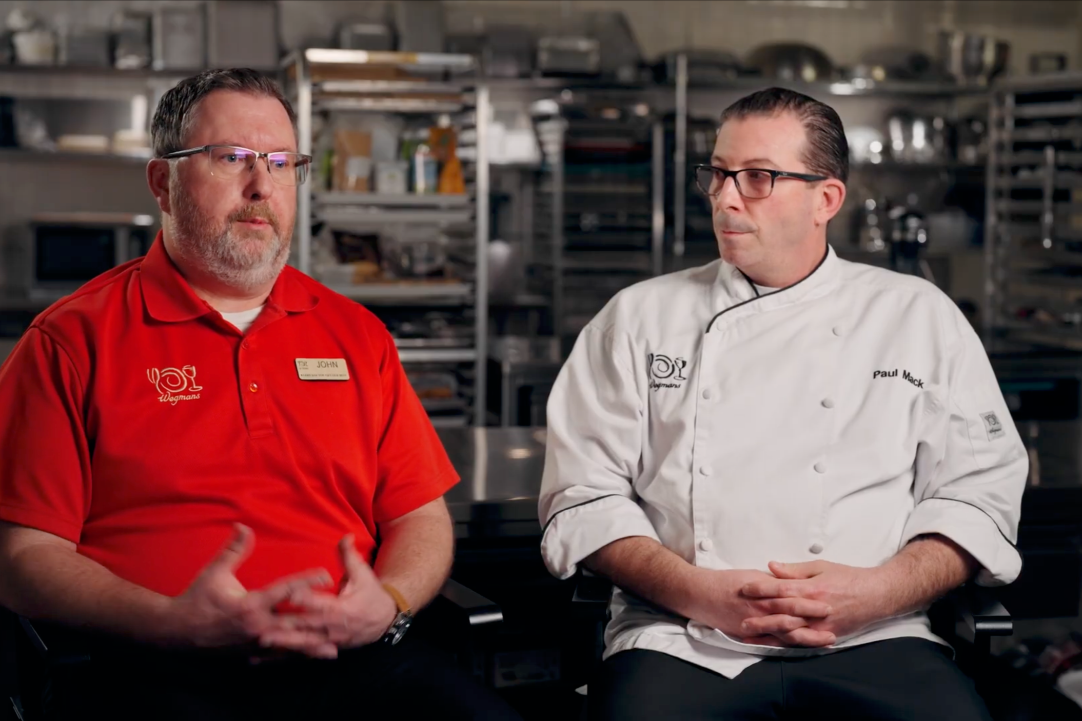 Two men sitting in a kitchen or restaurant setting, one wearing a red shirt with a name tag and the other wearing a white chef's coat, engaged in conversation.
