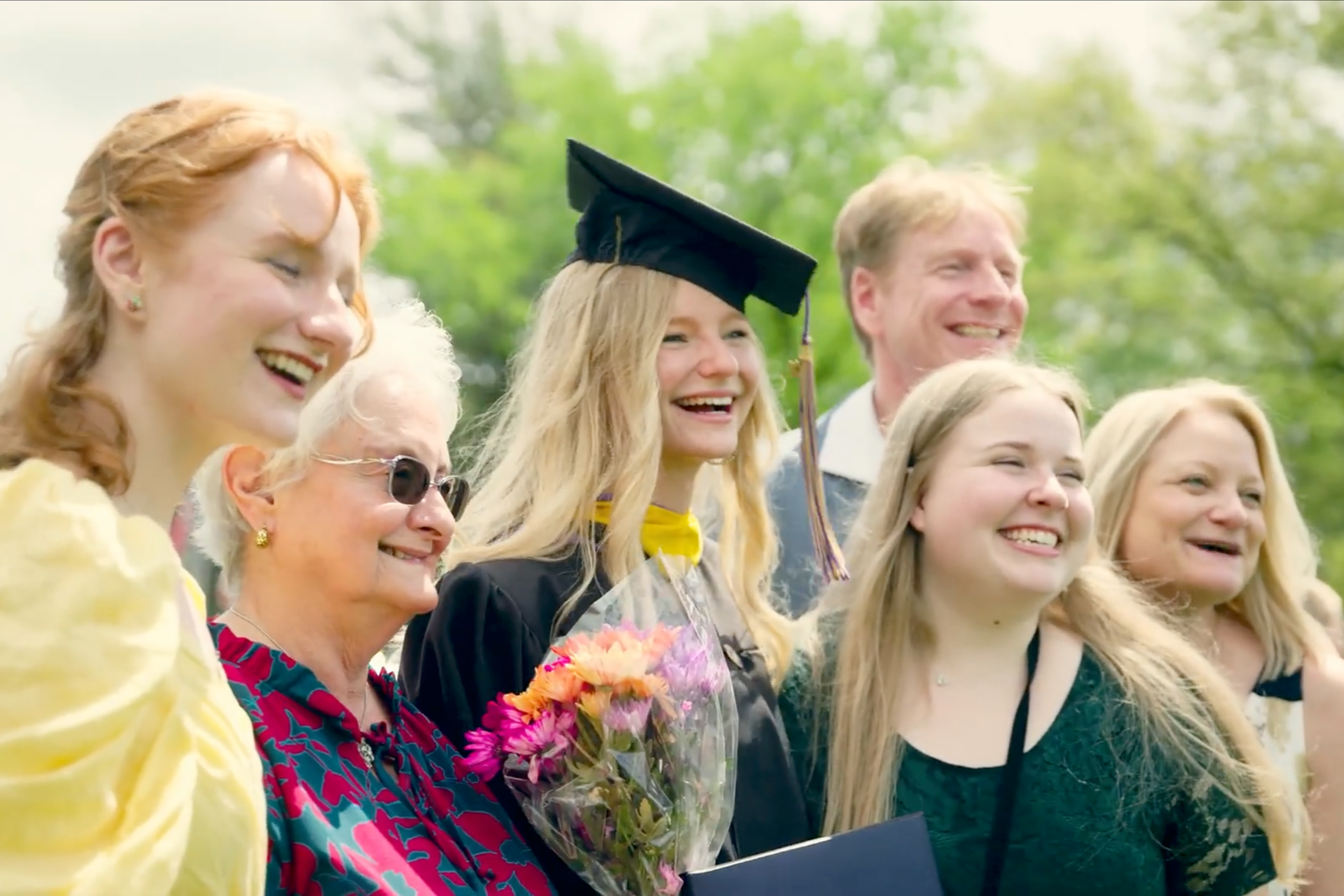 Group of people smiling outdoors, one woman in graduation cap and gown, holding a bouquet of flowers.
