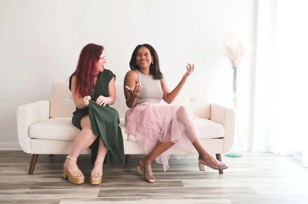 Two women sitting on a white sofa, smiling and talking, in a brightly lit room with a sheer curtain and a tall glass vase with dried plants in the background.