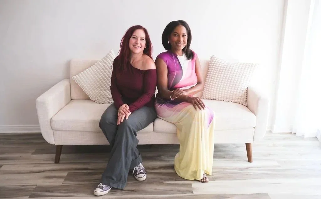 Two women sitting on a white couch in a bright room with white walls and wooden flooring, smiling at the camera.