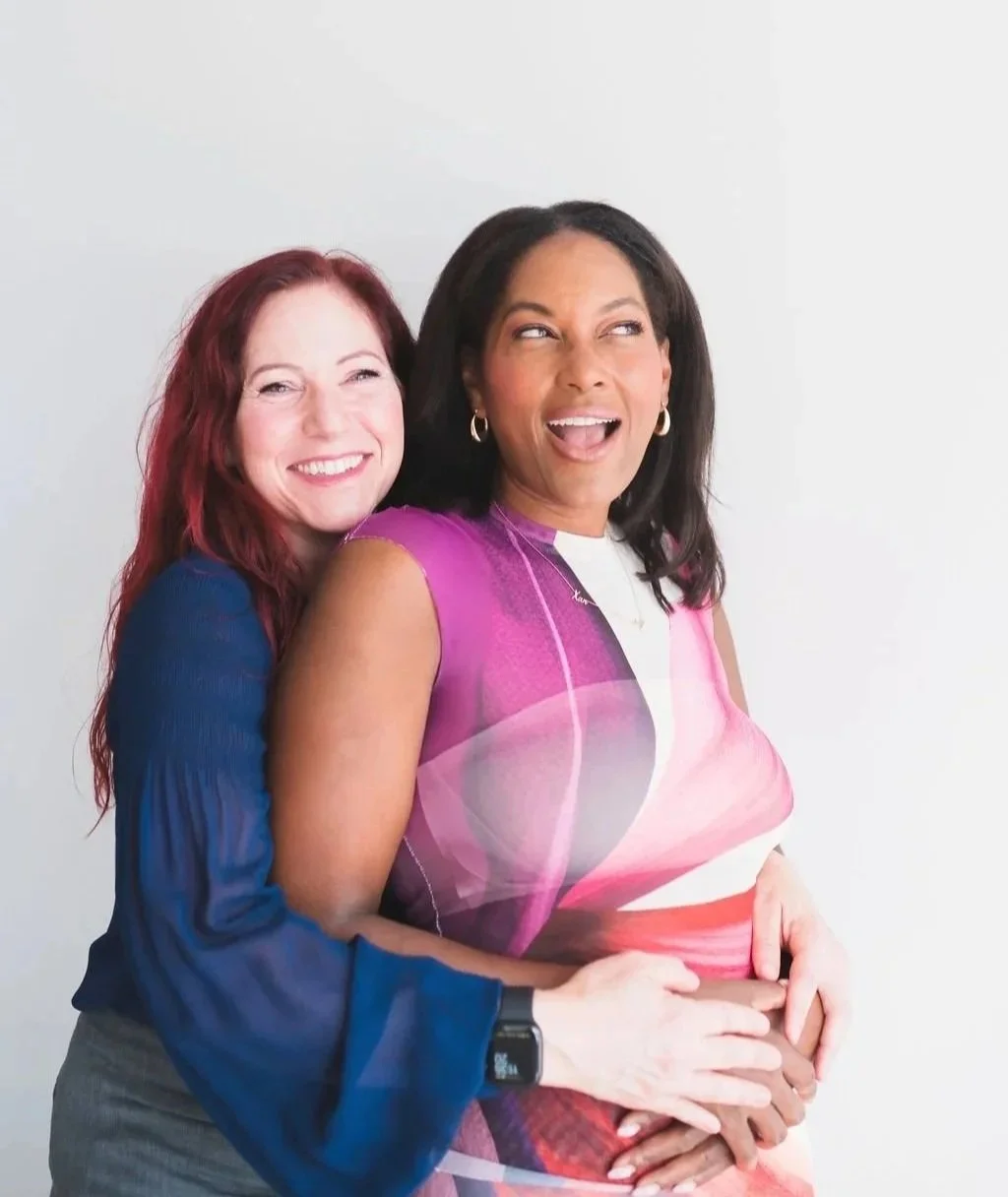 Two women smiling and embracing each other against a plain white background.