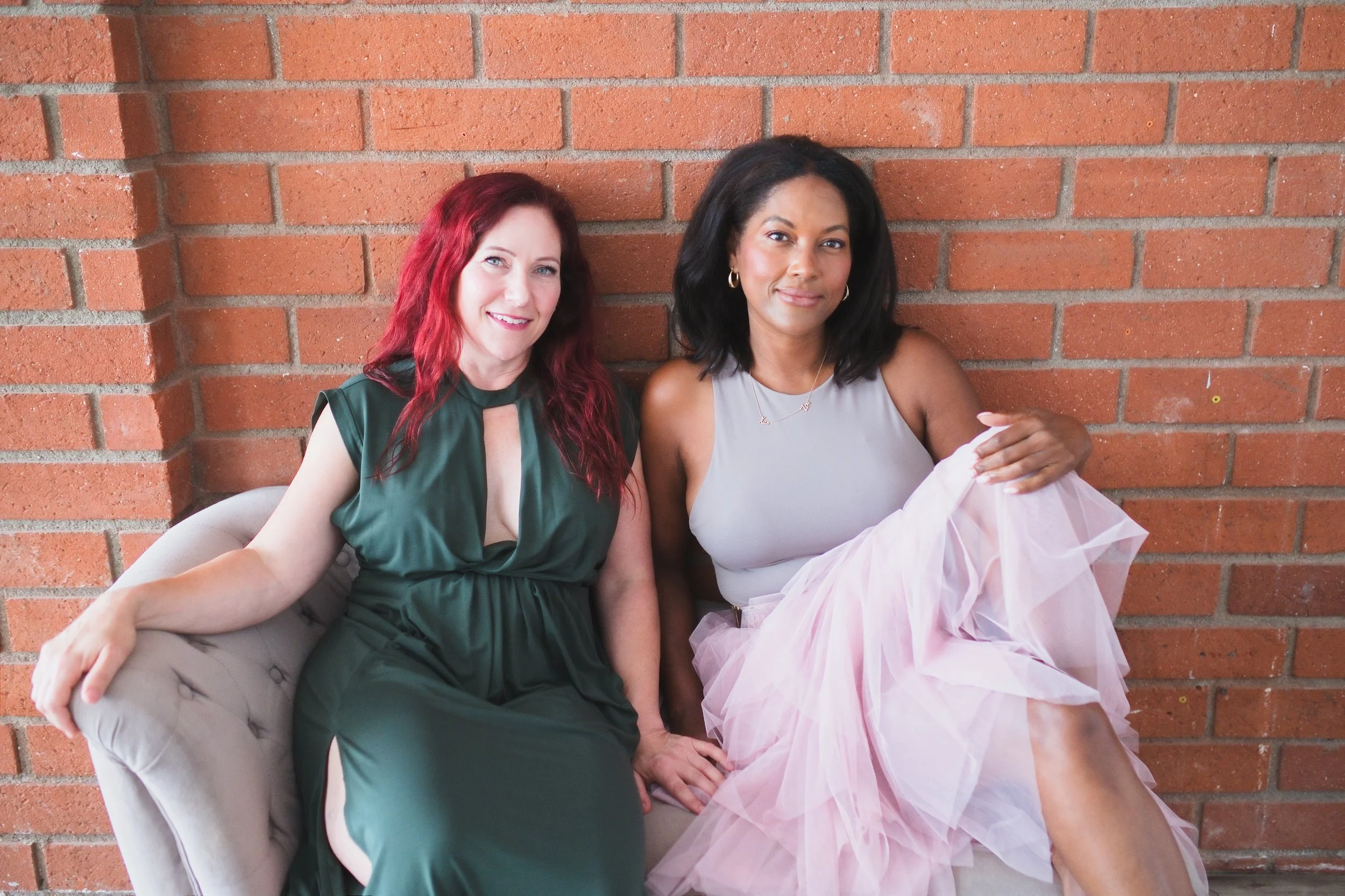Two women sitting on a beige tufted chair in front of a brick wall, smiling and looking at the camera. The woman on the left has red hair and is wearing a green dress. The woman on the right has black hair and is wearing a light-colored top and a pink tulle skirt.