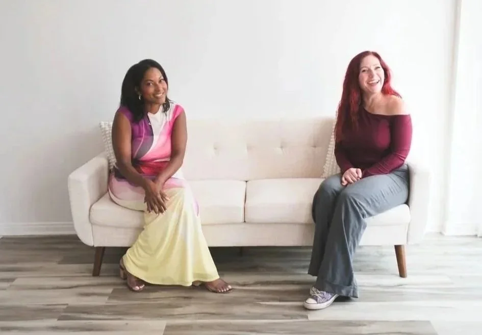 Two women sitting on a cream-colored couch in a bright, minimalistic room with gray wood flooring. The woman on the left is wearing a colorful dress and smiling, while the woman on the right has red hair, is wearing a burgundy off-the-shoulder top and gray pants, and is also smiling.
