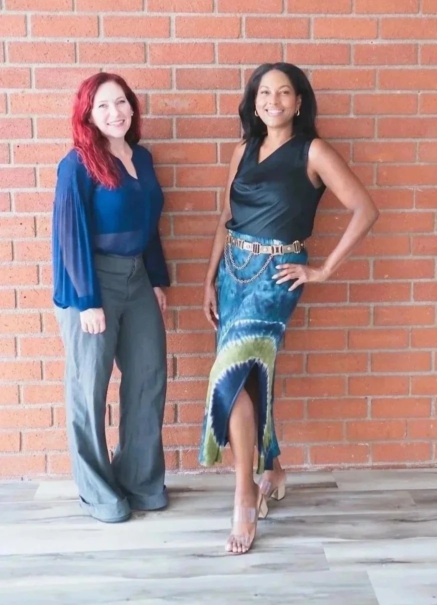 Two women standing in front of a red brick wall, smiling at the camera. The woman on the left has red hair and is wearing a blue blouse and gray pants. The woman on the right has black hair and is wearing a black sleeveless top, a colorful tie-dye skirt, and clear high heel shoes.