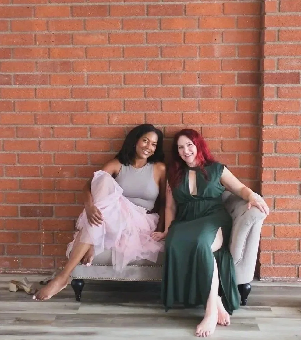 Two women sitting on a beige couch against a brick wall, smiling at the camera. The woman on the left has dark hair and is wearing a sleeveless gray top and a pink tutu, with her legs extended and toes pointed. The woman on the right has red hair, a green dress with a slit, and is barefoot, with her arm resting on the armrest.