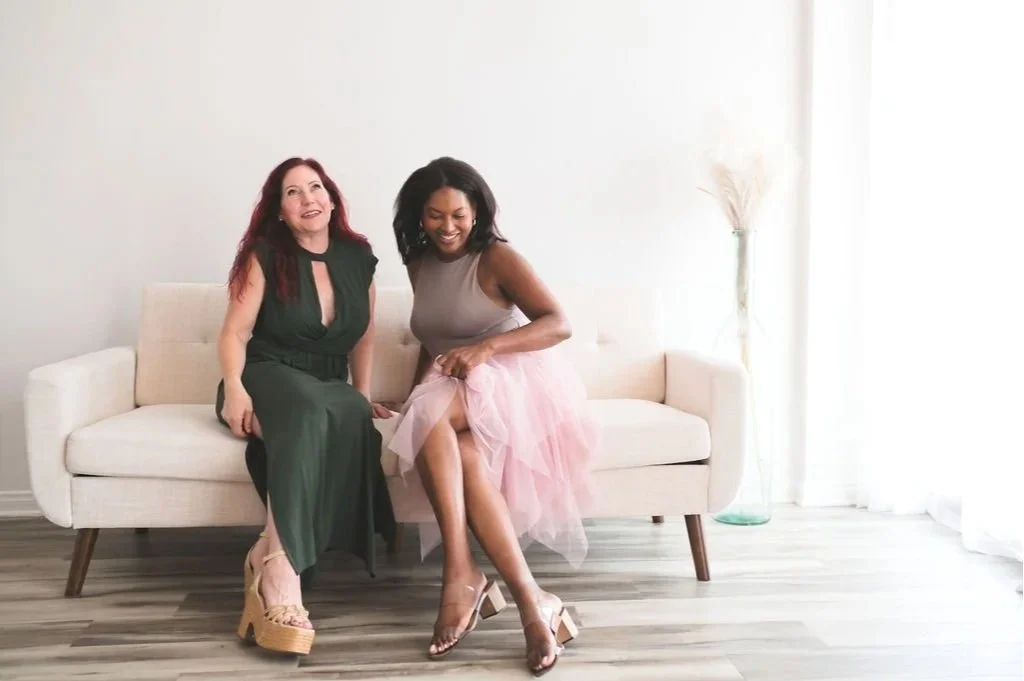 Two women sitting on a beige sofa, smiling, in a well-lit, minimalistic room with light-colored walls and wooden flooring.