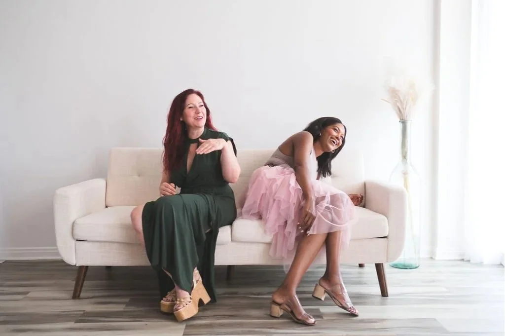 Two women sitting on a white sofa, laughing and enjoying each other's company in a well-lit room with a vase of dried flowers in the background.