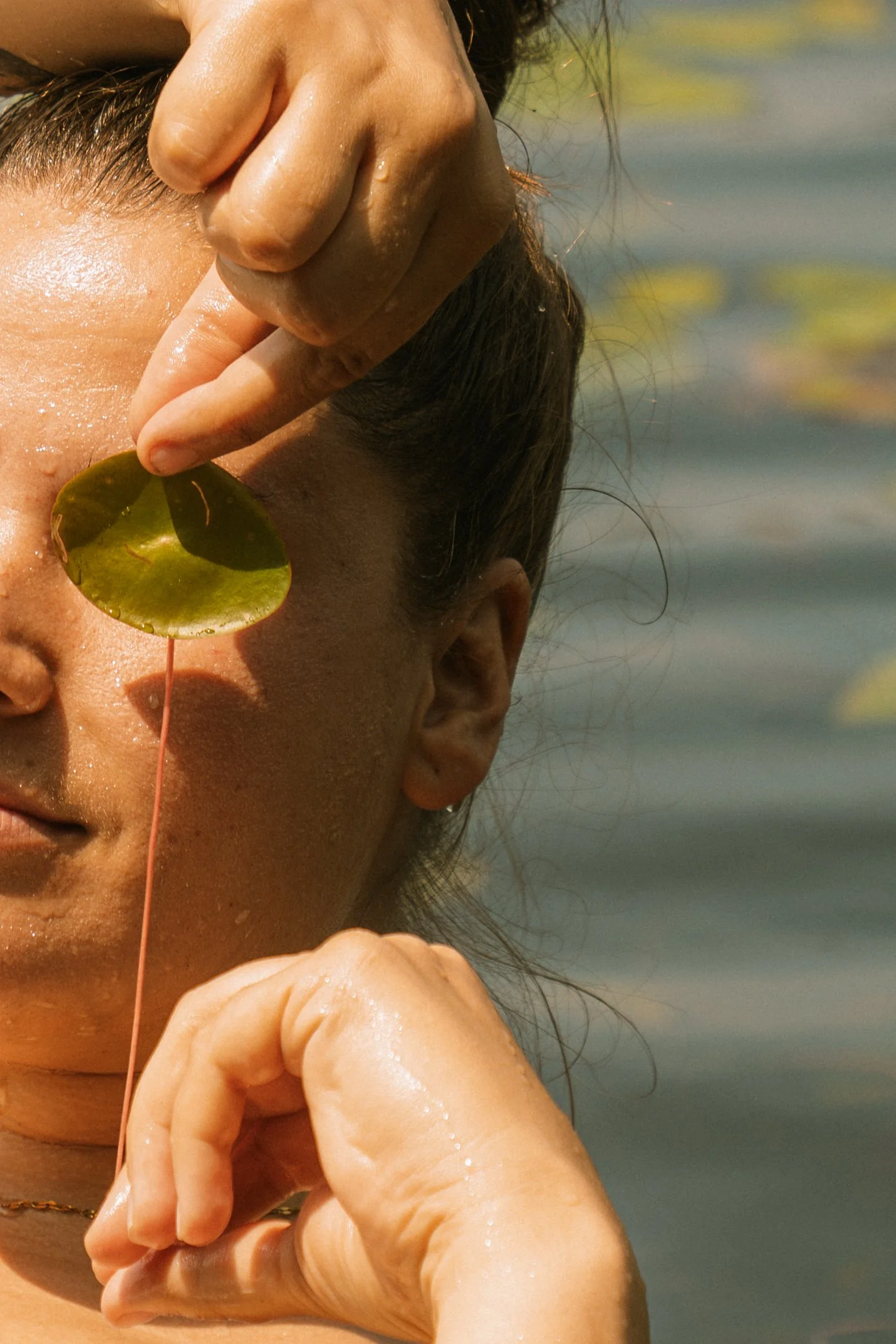 Portrait lifestyle spontané avec une feuille de nénuphar devant l’œil, capturé près de l’eau dans une ambiance naturelle et ludique.