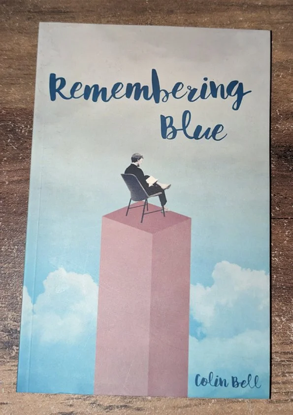 Book cover titled 'Remembering Blue' by Colin Bell, featuring an illustration of a person sitting on a chair atop a pink rectangular column with a cloudy sky background.