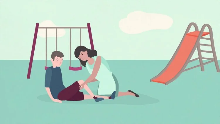 A girl helping a boy who is sitting on the ground near a playground swing set and slide.