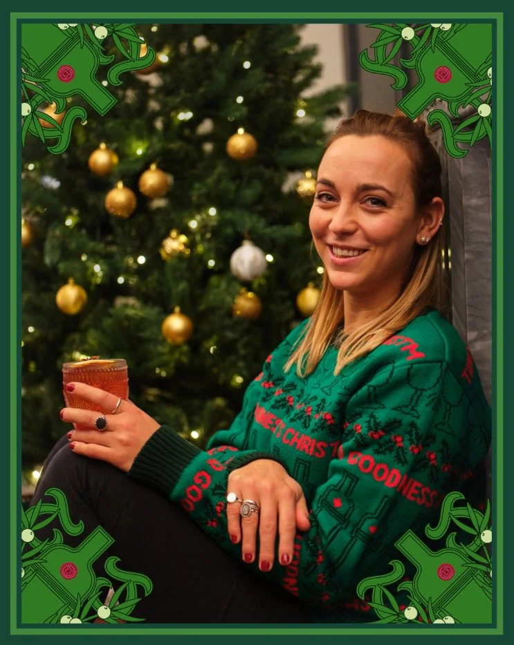 A woman smiling and sitting in front of a decorated Christmas tree with gold and white ornaments, holding a cup, wearing a Christmas sweater, with decorative border featuring holly and Christmas ornaments.