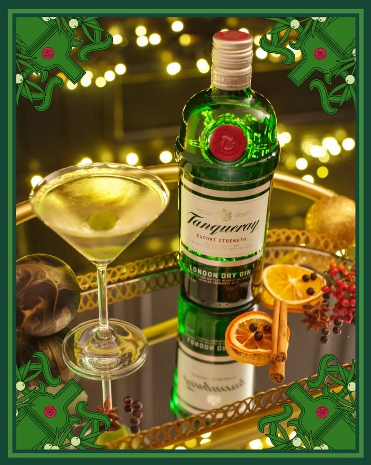 A bottle of Tanqueray London Dry Gin on a reflective glass surface with a martini glass containing a green olive, slices of dried orange, cinnamon sticks, and black peppercorns nearby, decorated with holiday elements and warm bokeh lights.