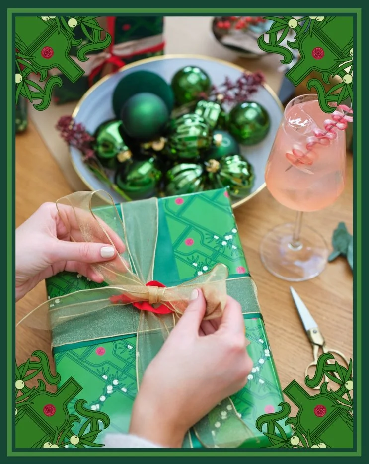 Person wrapping a Christmas present with green patterned wrapping paper and a gold ribbon, with a pair of scissors nearby, on a wooden table. In the background, a bowl of green Christmas ornaments and a glass of pink cocktail with a striped straw are