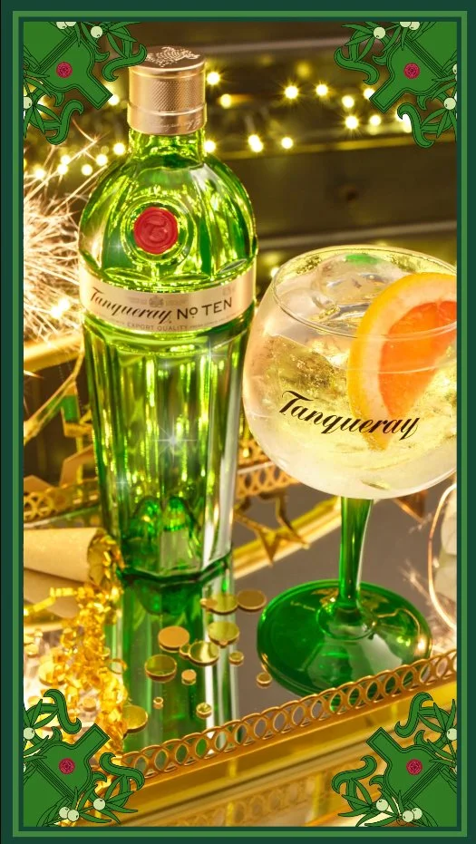 A bottle of Tanqueray gin and a large gin and tonic drink garnished with a slice of grapefruit, set on a reflective surface with festive lighting and gold decorations in the background.