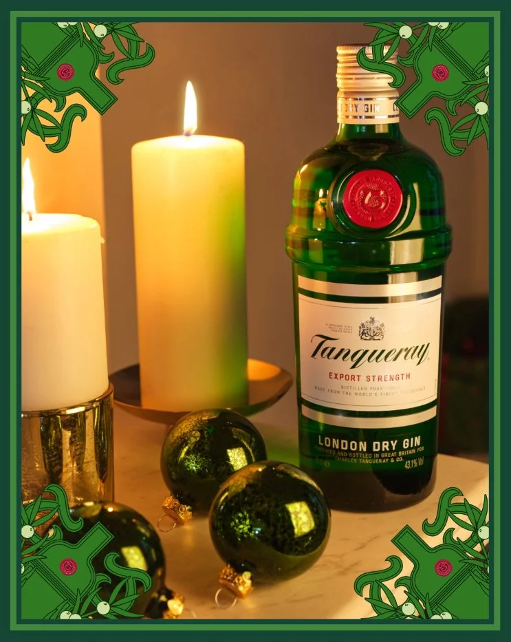 A photo of three candles, a bottle of Tanqueray London Dry Gin, and three green Christmas ornaments on a marble surface. The image has a festive border with holly and berries.