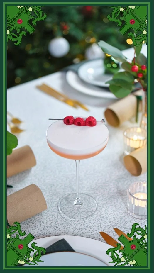A festive holiday table setting featuring a white cocktail with three red berries on top, surrounded by candles, green leaves, and Christmas decorations.