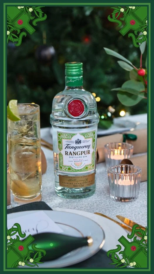 Bottle of Tanqueray Rangpur gin on a table next to a glass of gin and tonic with lime, with candles and Christmas decorations in the background.