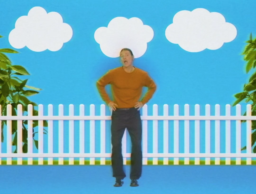 Person standing in front of a colorful backdrop featuring a blue sky, white clouds, green bushes, and a white picket fence.