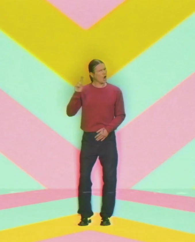 A person with long dark hair, wearing a red sweater and dark pants, stands against a colorful, abstract background with pastel yellow, pink, and aqua geometric shapes, making a hand gesture and facial expression.