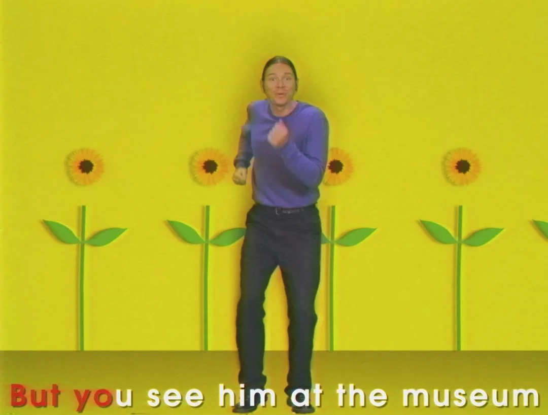 Person in a purple sweater standing in front of a yellow wall with sunflower decorations, with a caption that reads 'But you see him at the museum.'