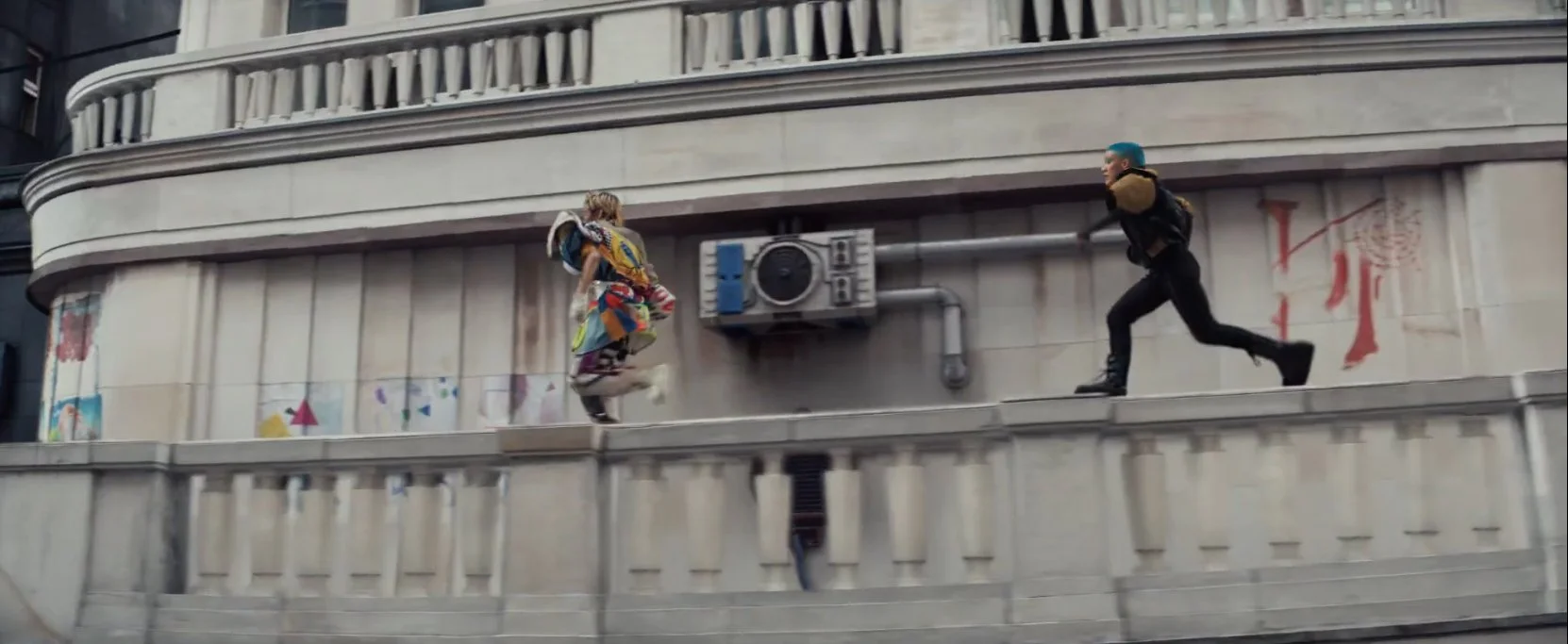 Two individuals running along a ledge outside a building with graffiti and an air conditioning unit.