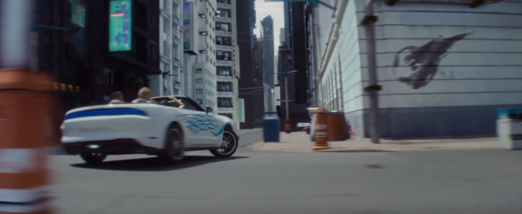 A white convertible car with blue racing stripes driving through a city street with tall buildings, street cones, and a mural of a horse on a wall.
