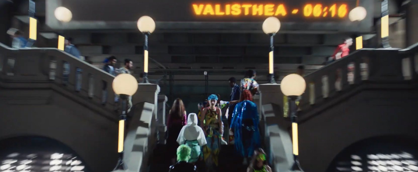 People walking up a staircase in a station or building, with an electronic display above showing the word 'VALISTEA' and a countdown timer reading 06:16. The scene is indoors with modern lighting and architectural features.