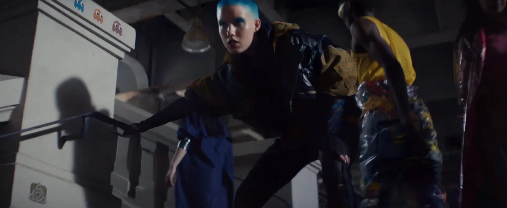 A person with short blue hair crouches on the ground in a dark room, surrounded by vending machines and other equipment, appearing distressed or intense.