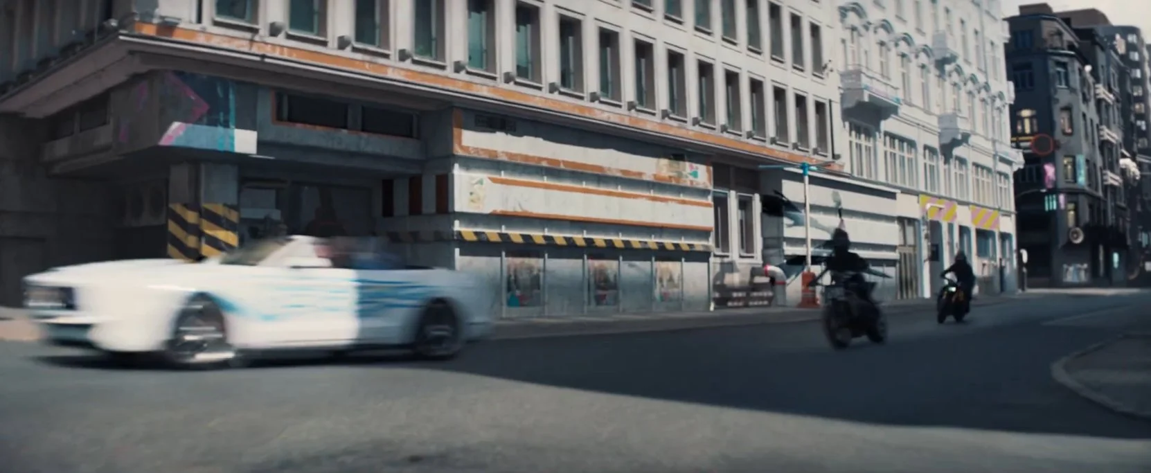 A white police car with blue stripes driving on an urban street with buildings, sidewalk, and people on motorcycles and walking.