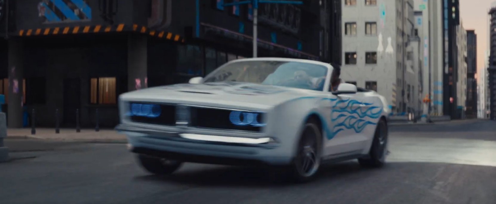 A white convertible car with blue flame decals driving through city streets during dusk or dawn, with tall buildings in the background.