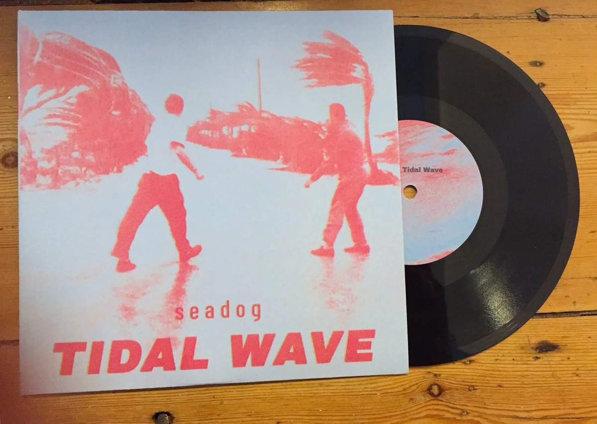 Album cover titled "Tidal Wave" by Seadog, featuring a pink and blue tinted image of two people walking near a boat on a body of water, with a vinyl record partially visible.