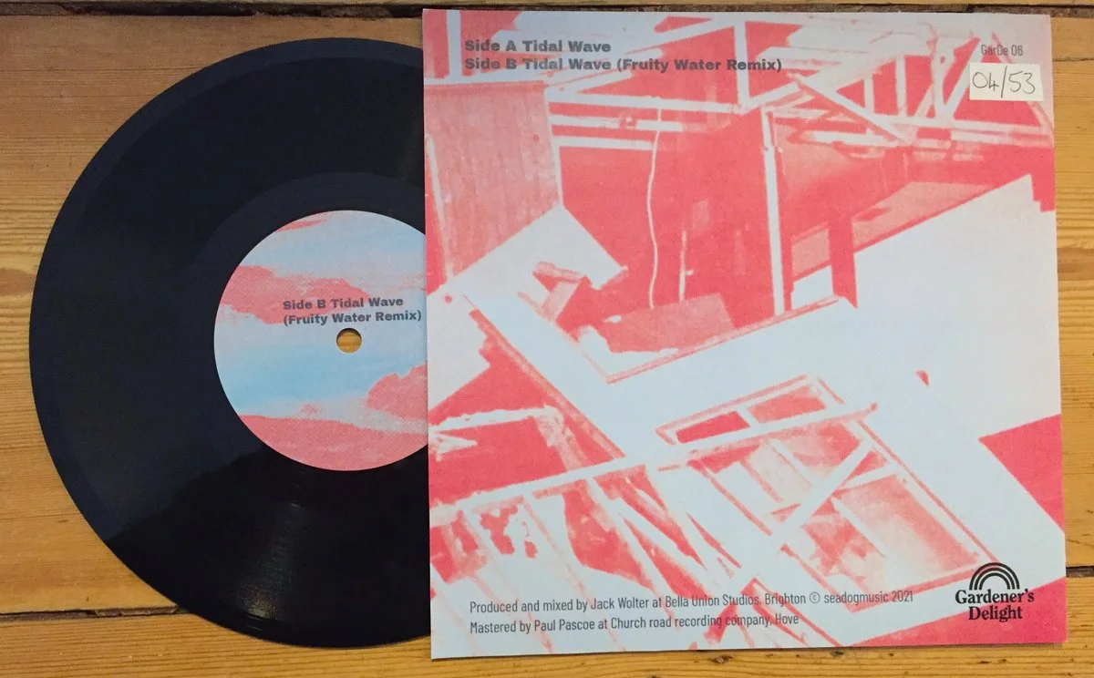 A vinyl record and its cover sleeve on a wooden surface. The cover has abstract red and blue artwork and text indicating it contains Tidal Wave (Fruity Water Remix) on Side A and Side B. The label on the vinyl reads 'Side B Tidal Wave (Fruity Water R
