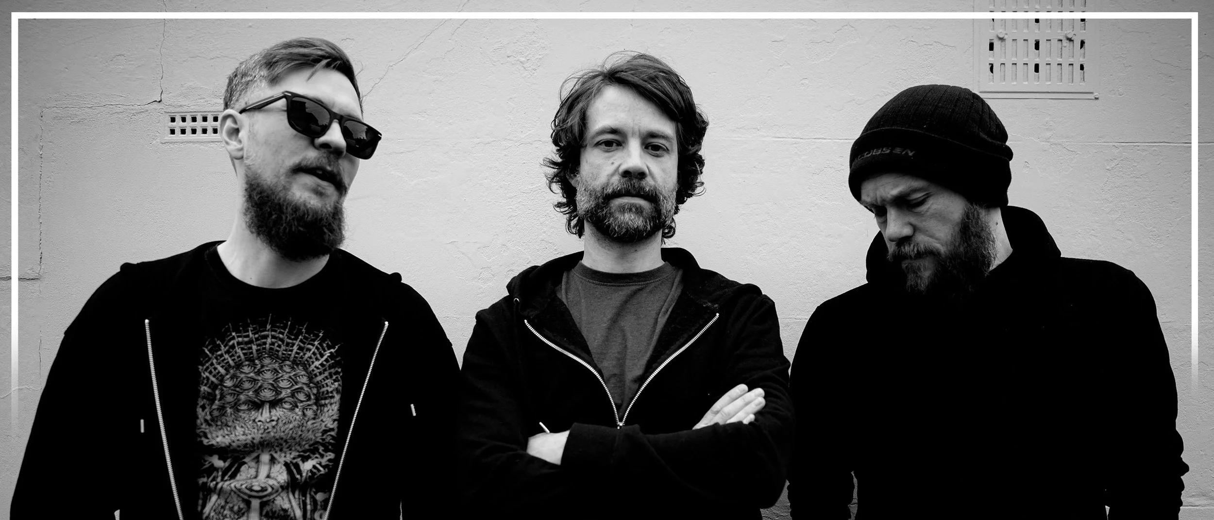Black and white photo of three men standing against a plain wall, with the man on the left wearing sunglasses and a graphic T-shirt, the man in the middle with crossed arms and a jacket, and the man on the right with a beanie and hoodie, all with beards.