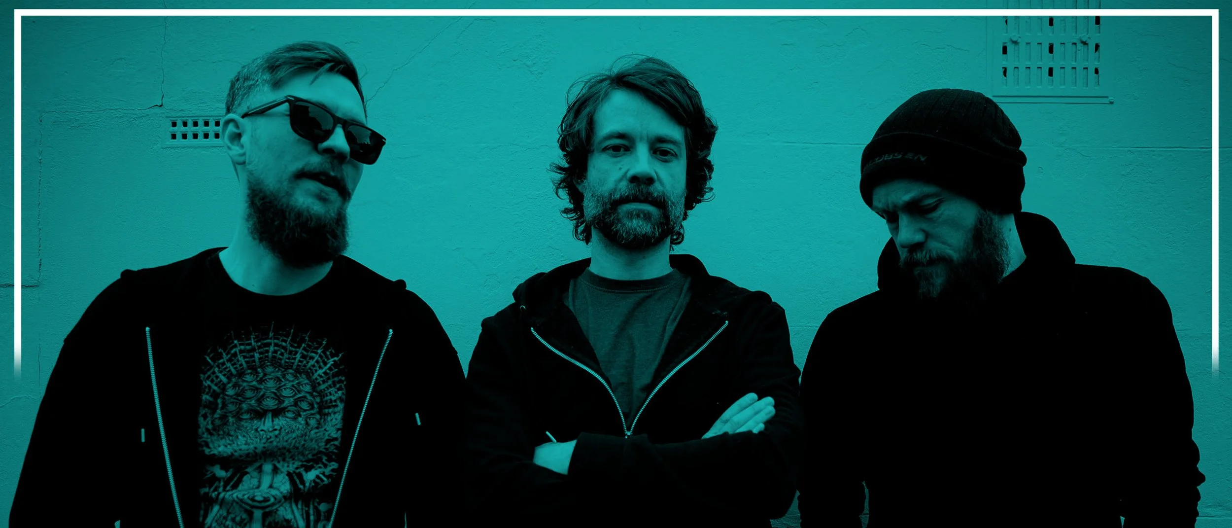 Three men standing against a plain wall, with the middle man crossing his arms and the others looking down or away, all wearing casual dark clothing.