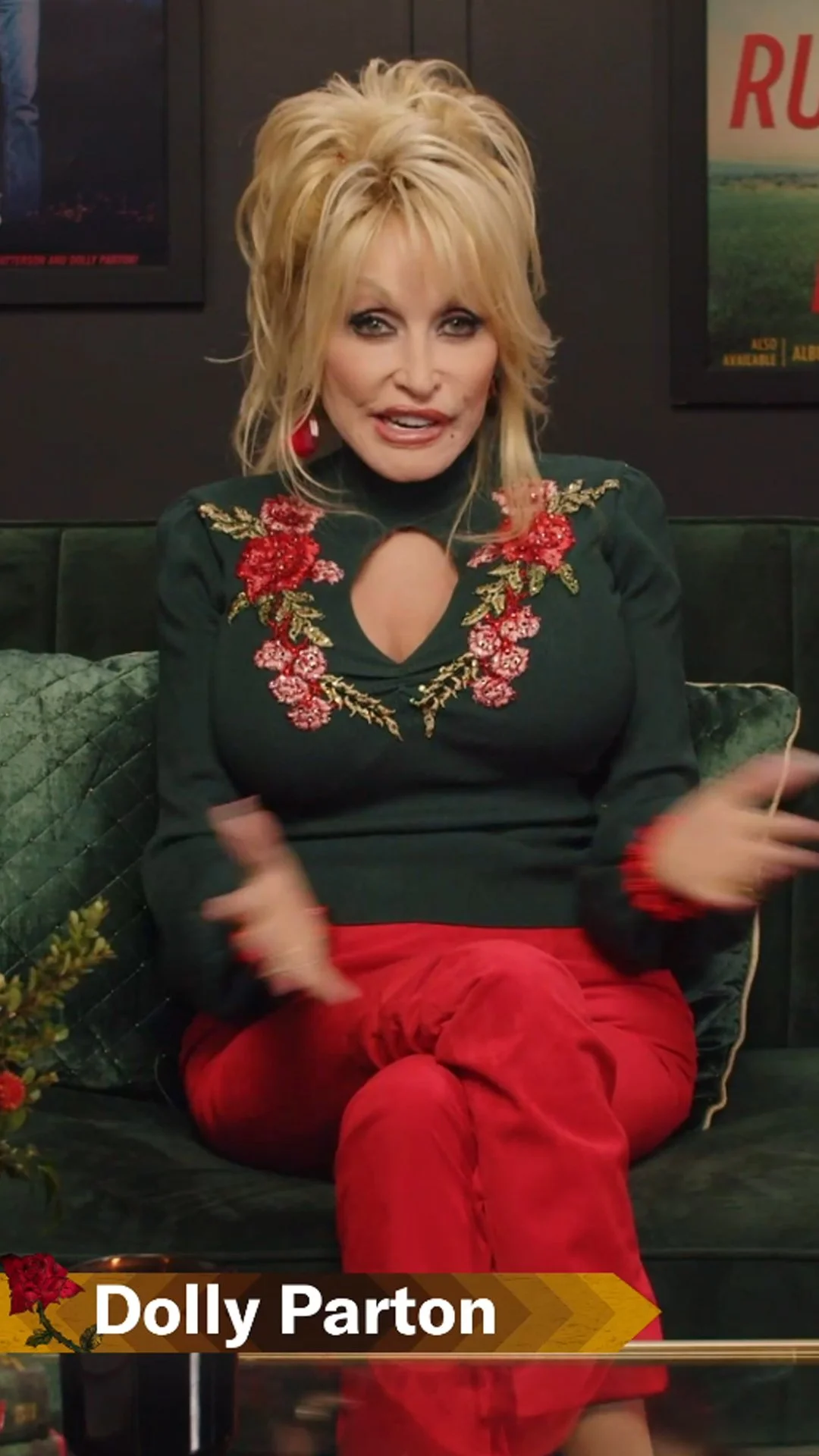 A blonde woman wearing a black top with red floral embroidery and red pants, sitting on a green couch, speaking, with framed posters on the wall behind her. The text overlay says 'Dolly Parton'.