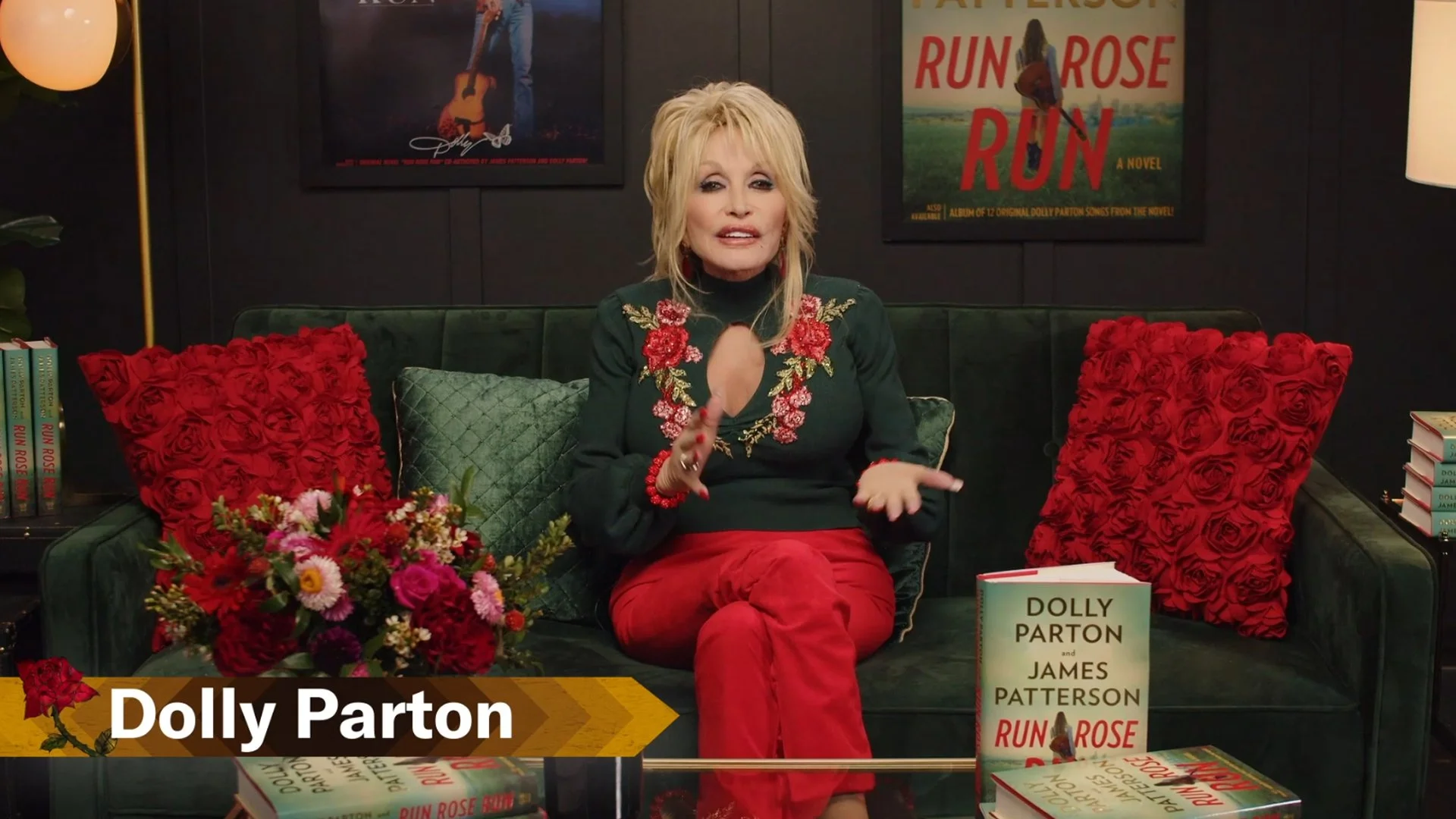 Author Dolly Parton sitting on a green velvet sofa with red and green pillows, surrounded by books and flowers, with posters of her book 'Run Rose Run' on the dark wall behind her.