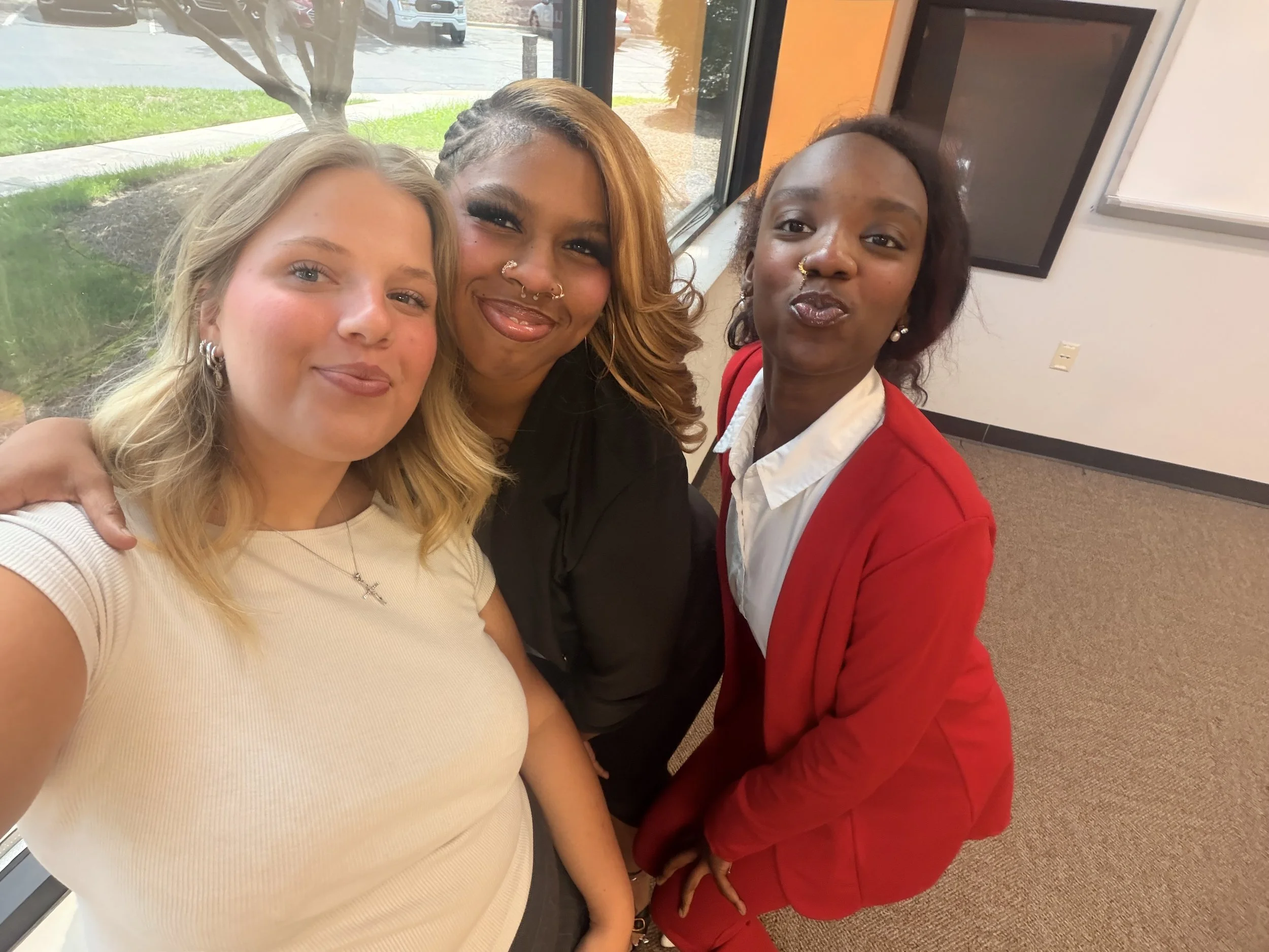 Three women taking a selfie in front of a large window. The woman on the left has blonde hair and fair skin, wearing a light-colored top. The woman in the middle has light brown skin, long brown hair with a hair braid on the side, and multiple facial piercings, wearing a black top. The woman on the right has dark skin, curly dark hair, and is wearing a white shirt with a red blazer. They are inside a building, with a parking lot and a tree visible outside.