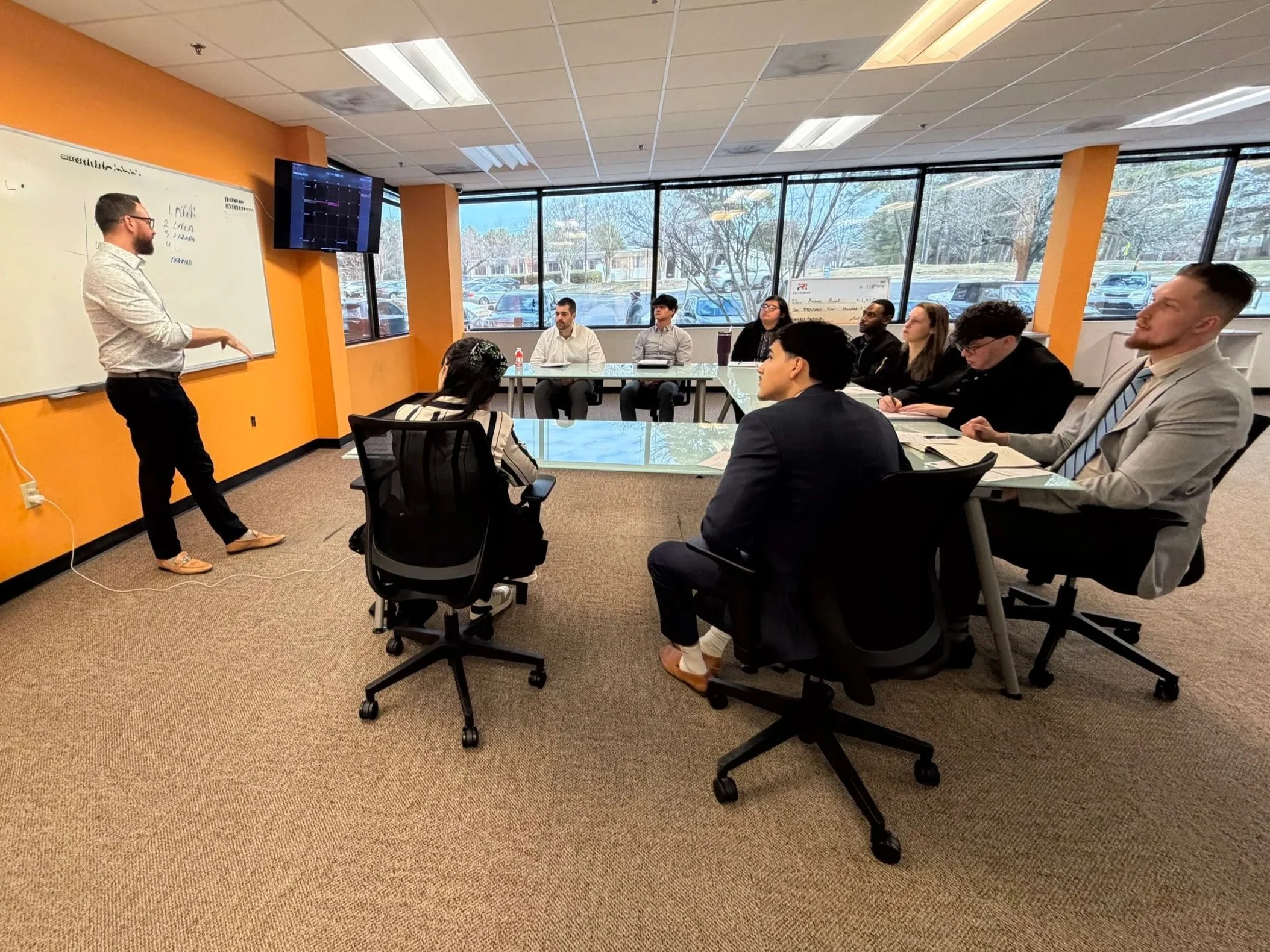 A man is giving a presentation in a conference room with large windows. He is standing by a whiteboard and talking to an audience seated around a large table. The room has orange walls, a hanging television screen, and a view of parked cars and trees outside.