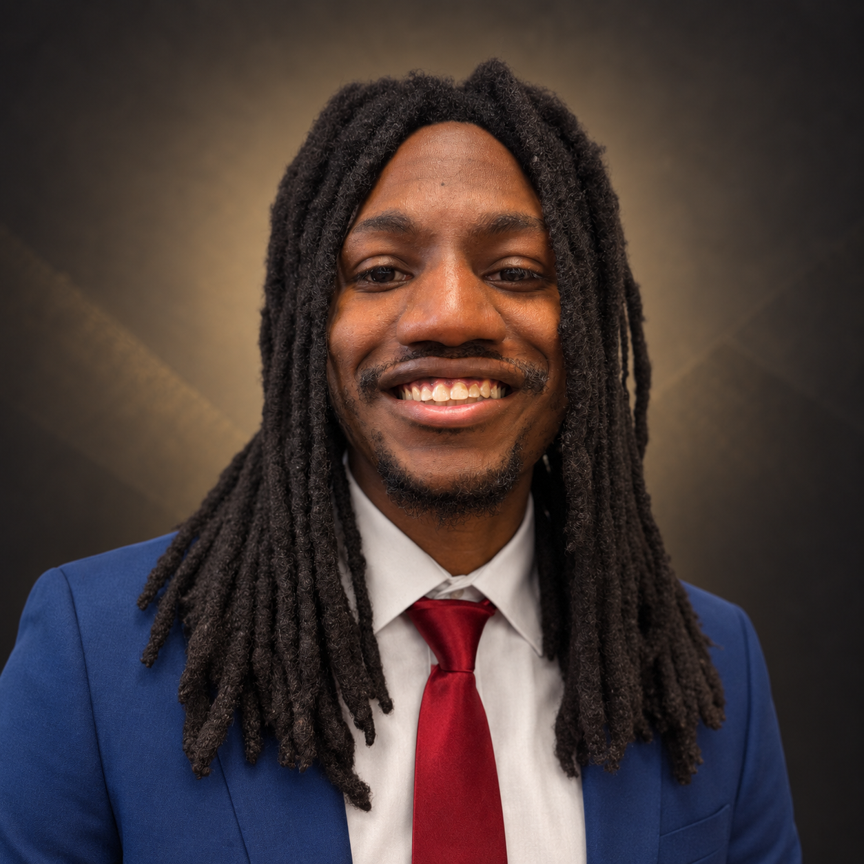 A smiling man with long dreadlocks, wearing a blue suit, white shirt, and red tie, against a dark, textured background.
