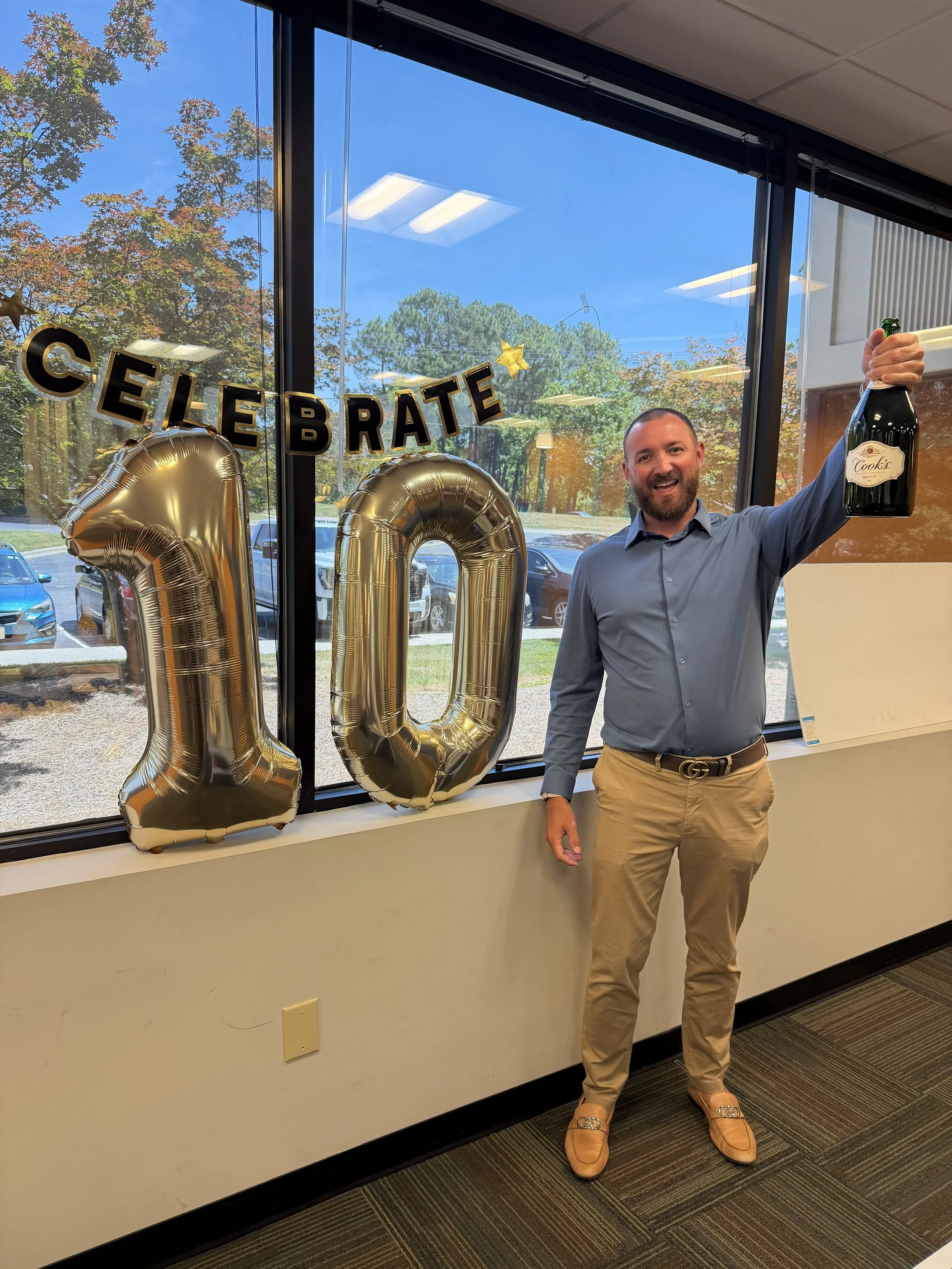 A man smiling and holding a bottle of sparkling wine standing next to gold balloon numbers forming '10' during a celebration event with 'Celebrate' banner and a window showing trees and parked cars outside.