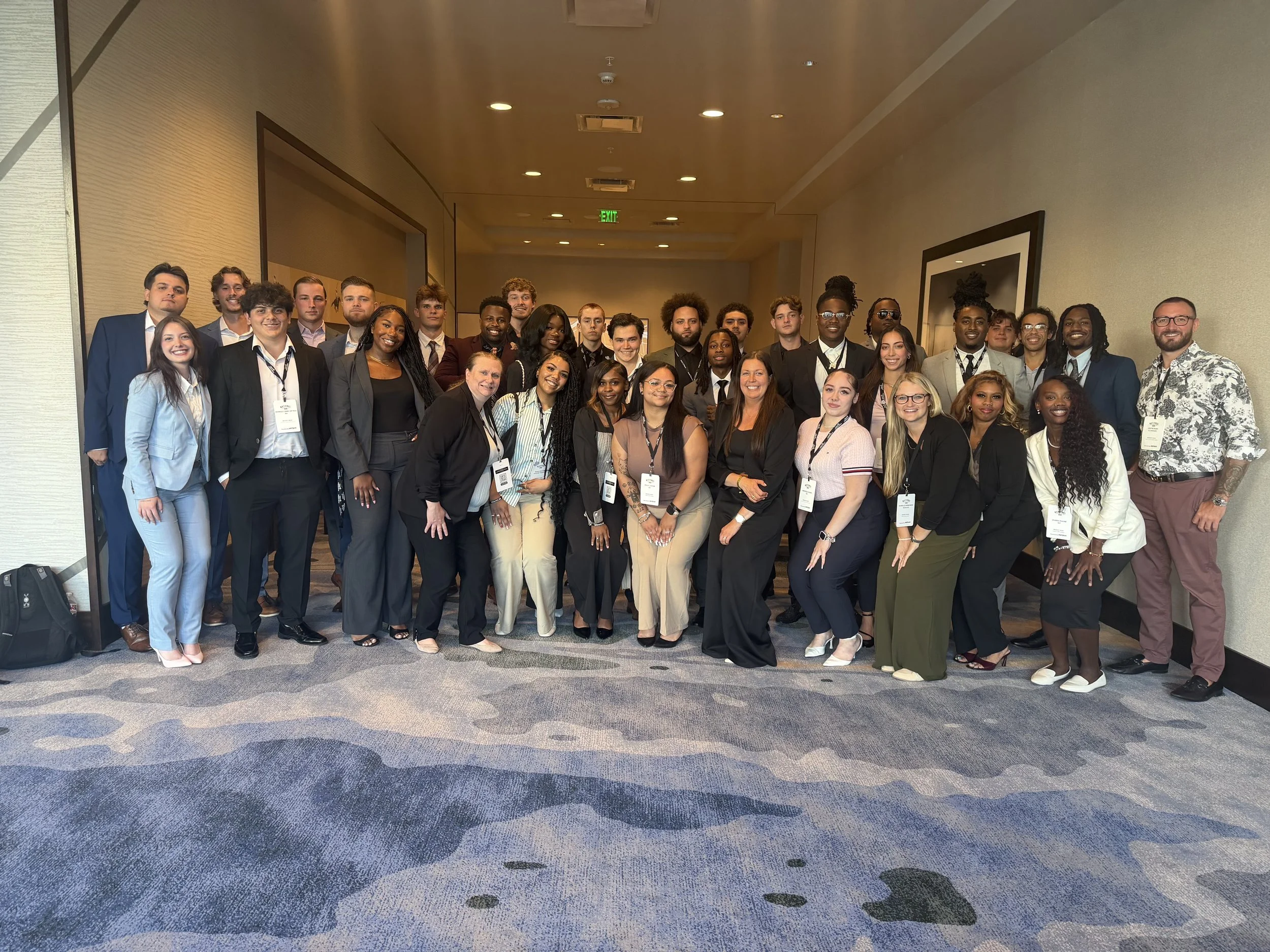 A large group of diverse people in professional attire standing together in a hallway, posing for a group photo at a conference or event.