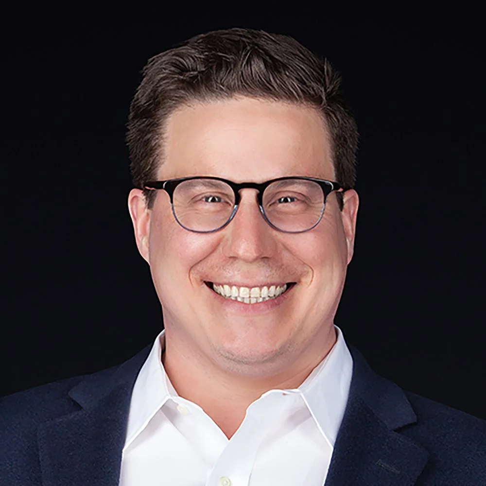 Headshot of a smiling man with glasses, wearing a white shirt and navy blazer, against a black background.