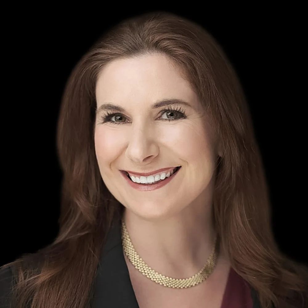 Close-up portrait of a woman with long brown hair, smiling, wearing a black blazer and a gold necklace, against a black background.
