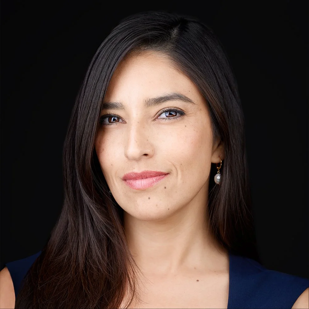 A woman with long dark brown hair, blue eyes, light skin, and gold earrings with pearls, smiling against a black background.